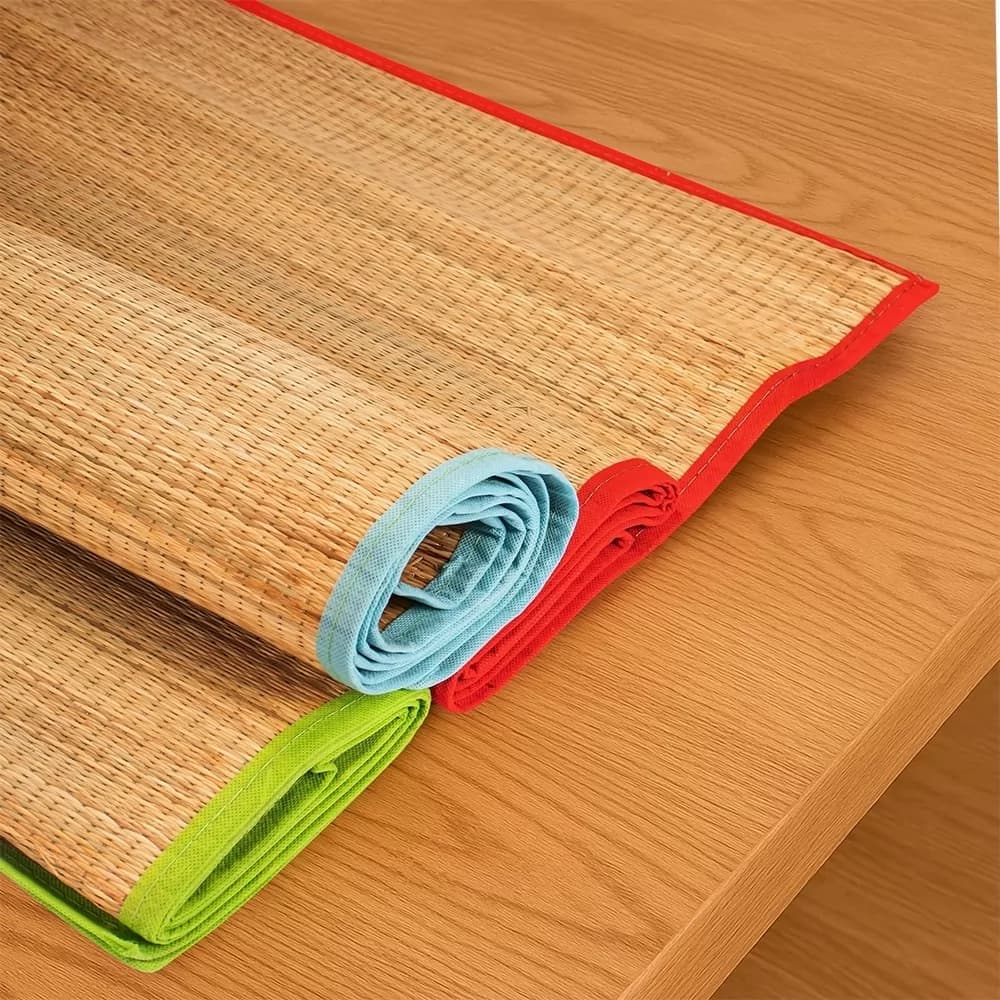 Esteiras de palha natural enroladas com bordas de tecido nas cores azul, verde e vermelho sobre mesa de madeira