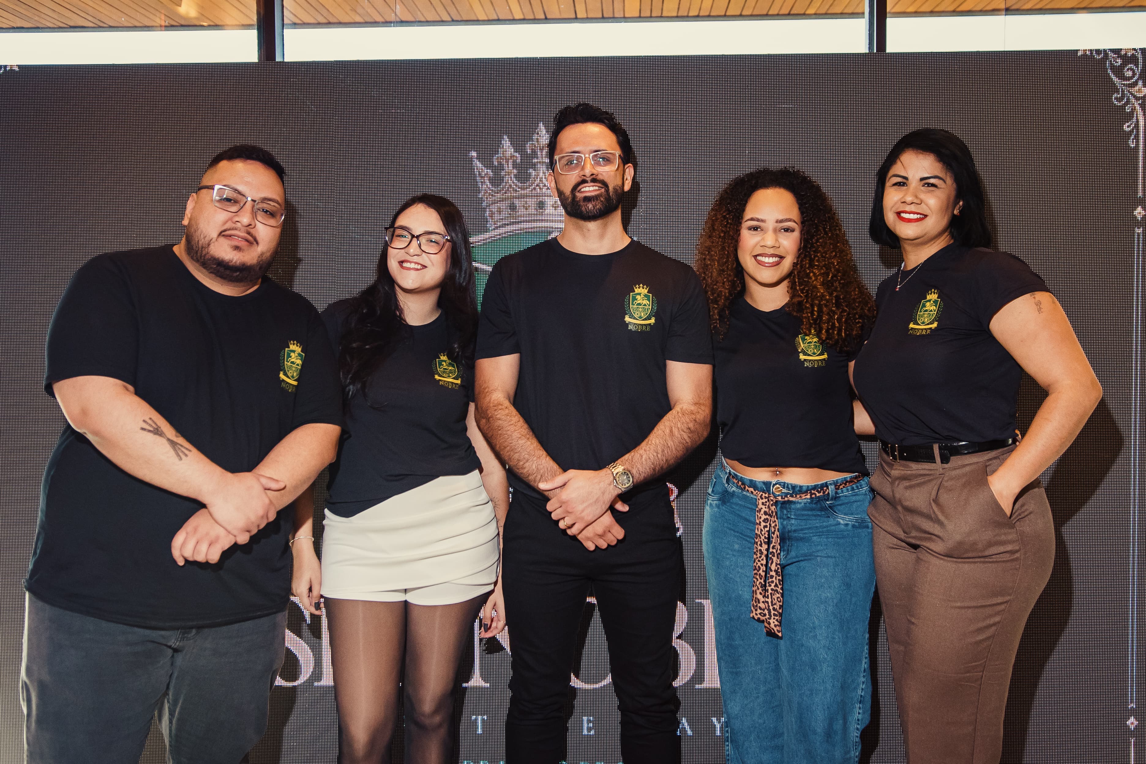 Equipe da Nobre Brindes reunida, com camisetas da marca, diante de um painel do evento.